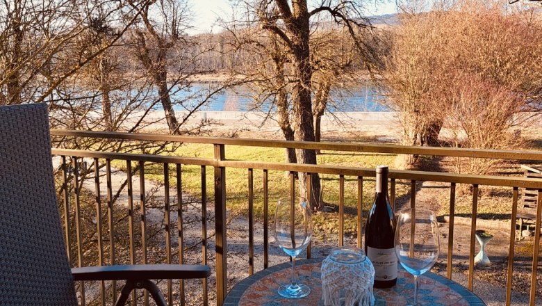 Balcony with table, two wine glasses and wine bottle, view of trees and river.