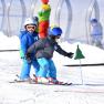 Two children in ski equipment skiing in the snow, accompanied by an adult in the background.
