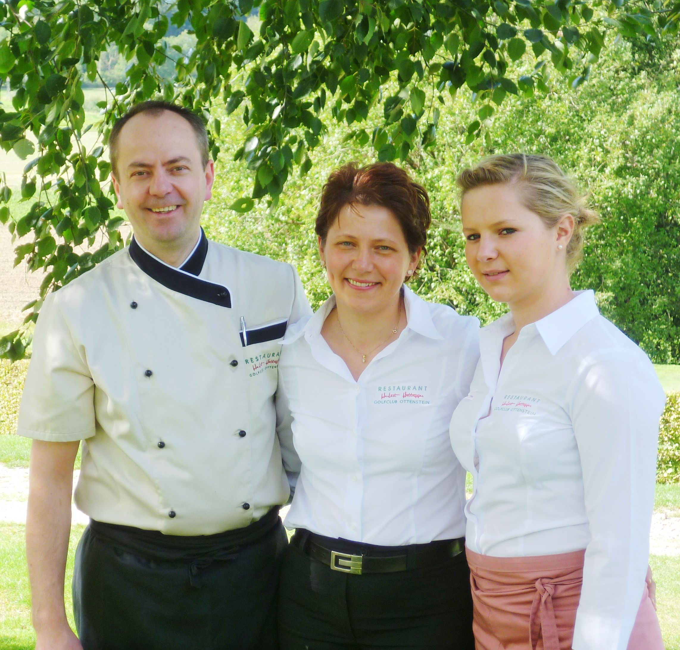 Three people in restaurant uniforms stand smiling under a tree.