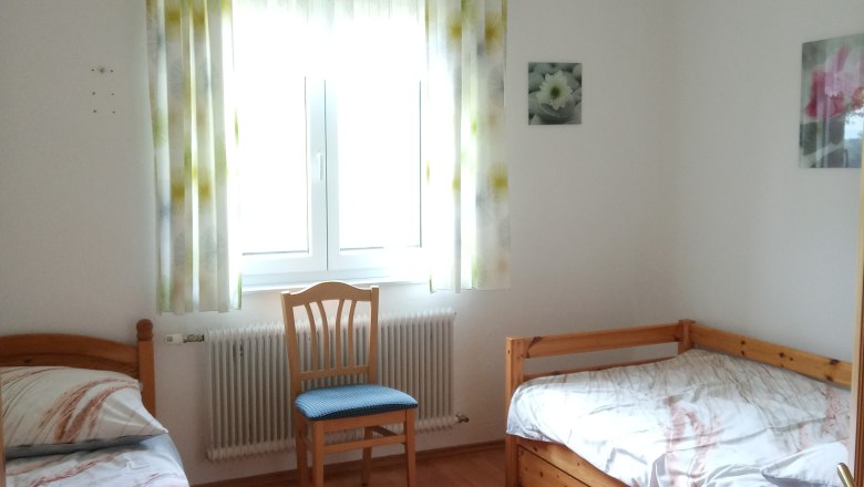 A simply furnished bedroom with two single beds, a chair and a window with curtains.