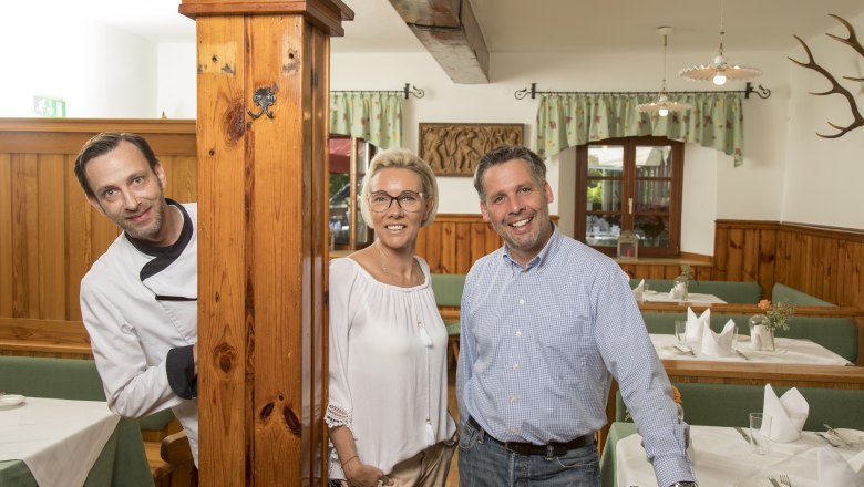 Three people in a rustic restaurant, one in chef's clothes, two in casual clothes, smiling at the camera.