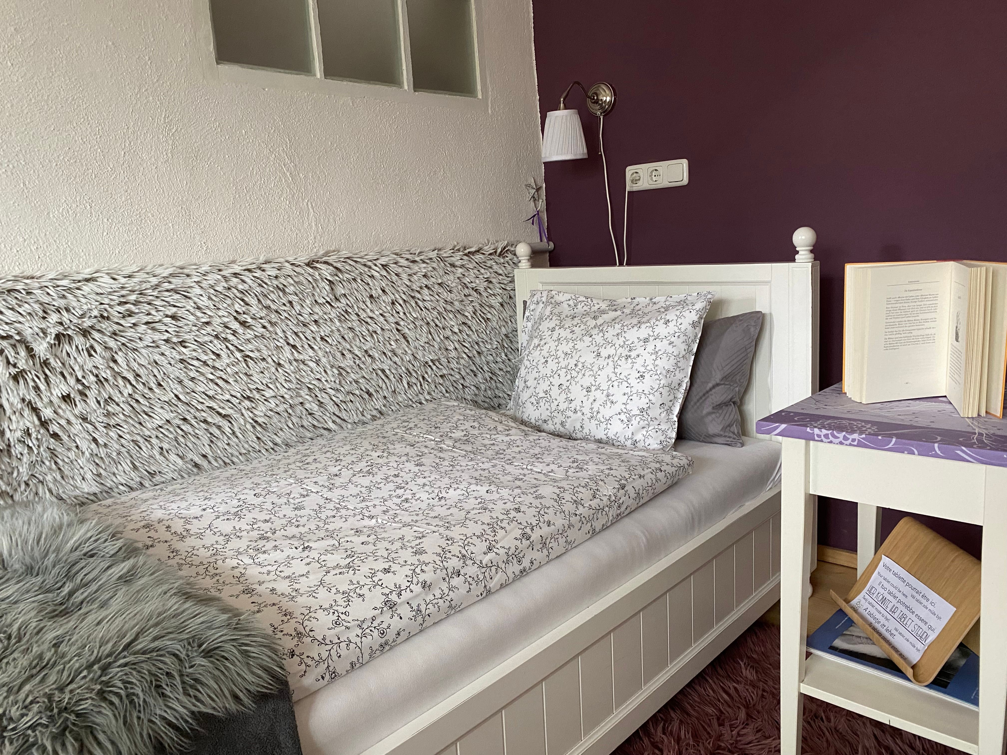 Cozy bedroom with single bed, purple wall, bedside table with book and lamp.