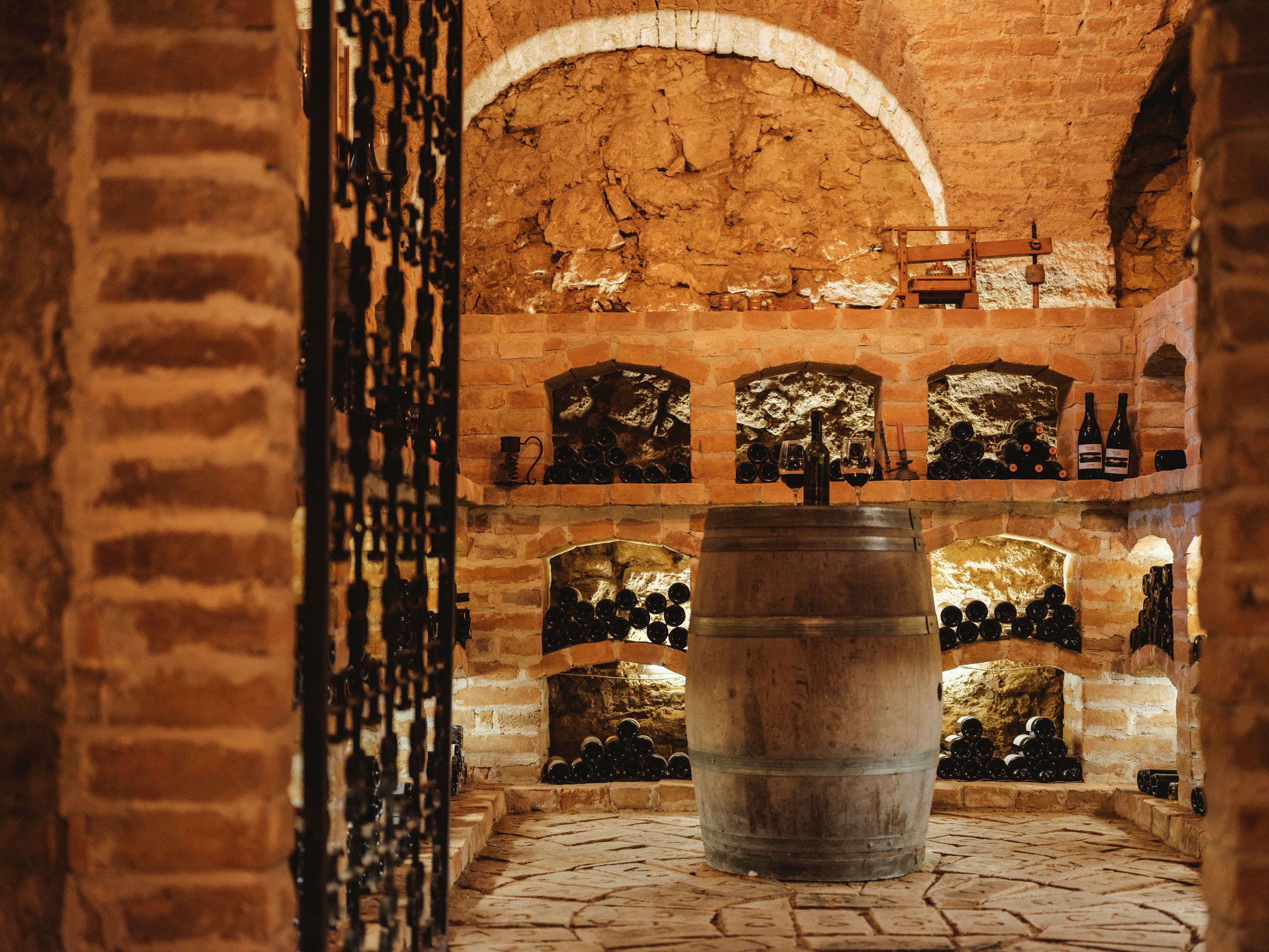 A rustic wine cellar with brick walls, wine racks and a wooden barrel in the middle.