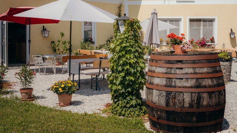 A cozy guest garden with parasols, wooden tables and a large wooden barrel, surrounded by plants and flowers.