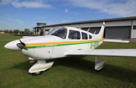 A small airplane, Piper Cherokee Archer II, stands on a green field in front of a hangar and a control tower.