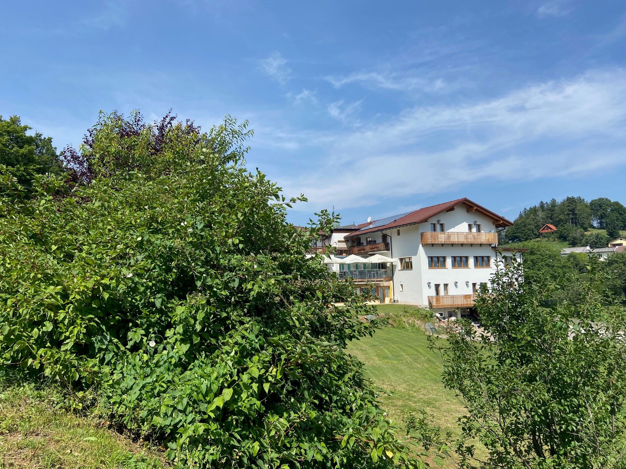 A white house with a red roof, surrounded by green trees and meadows, under a blue sky.