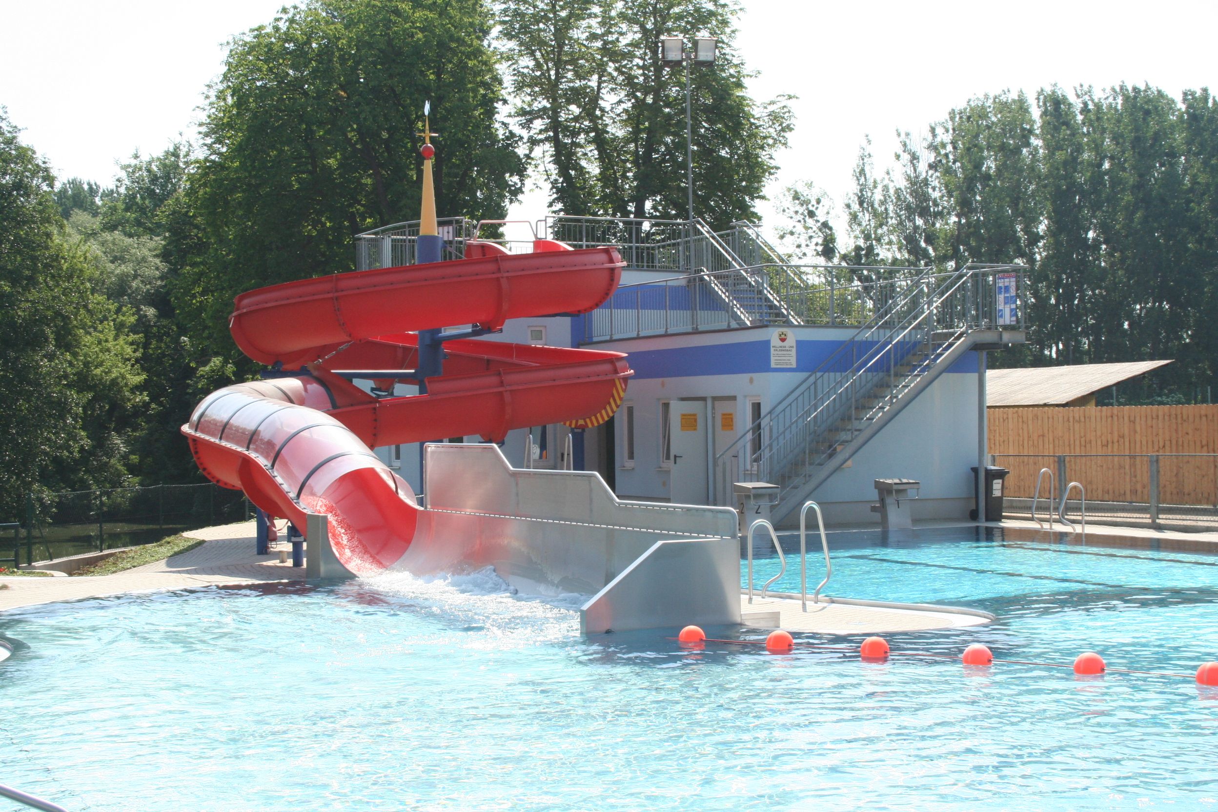 Red water slide in an outdoor pool with pool and trees in the background.