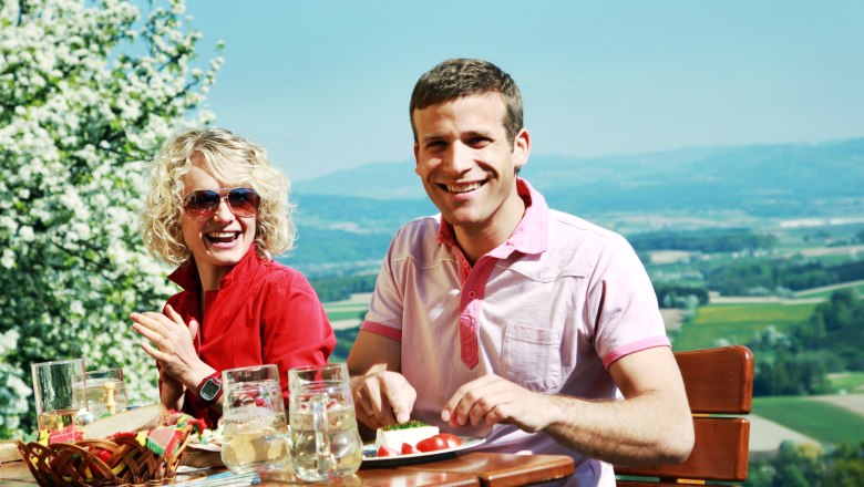 Two people sitting at an outdoor table with drinks and food, a landscape in the background.