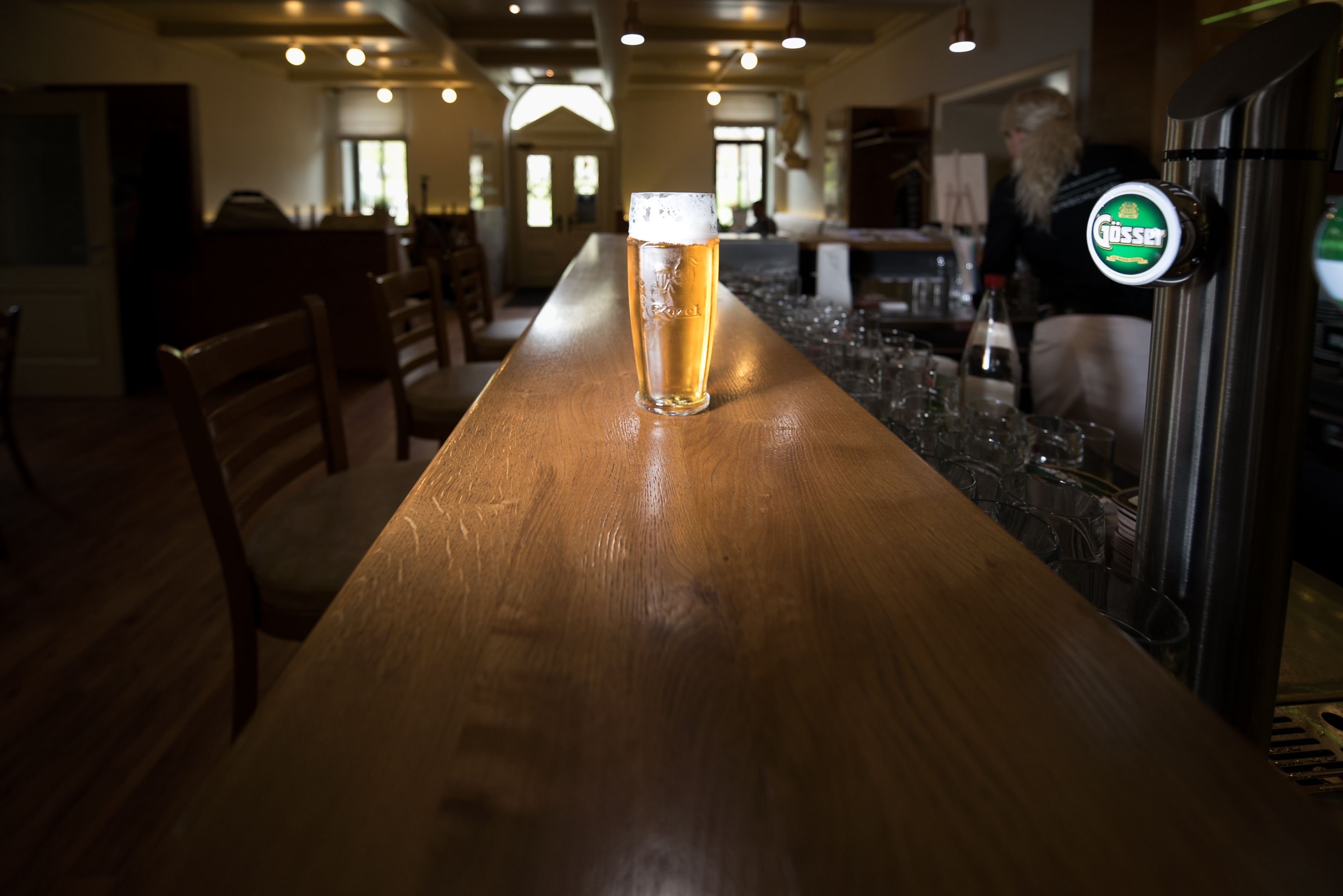 A glass of beer sits on a long wooden counter in a bar with empty chairs and a person in the background.