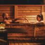 Two people in a wooden bathtub outdoors, clinking glasses of wine.