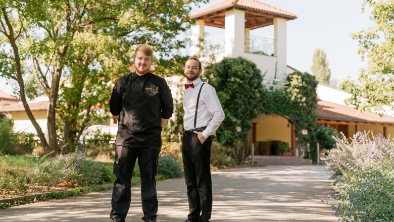 Two men in work clothes are standing in front of a building with a garden.