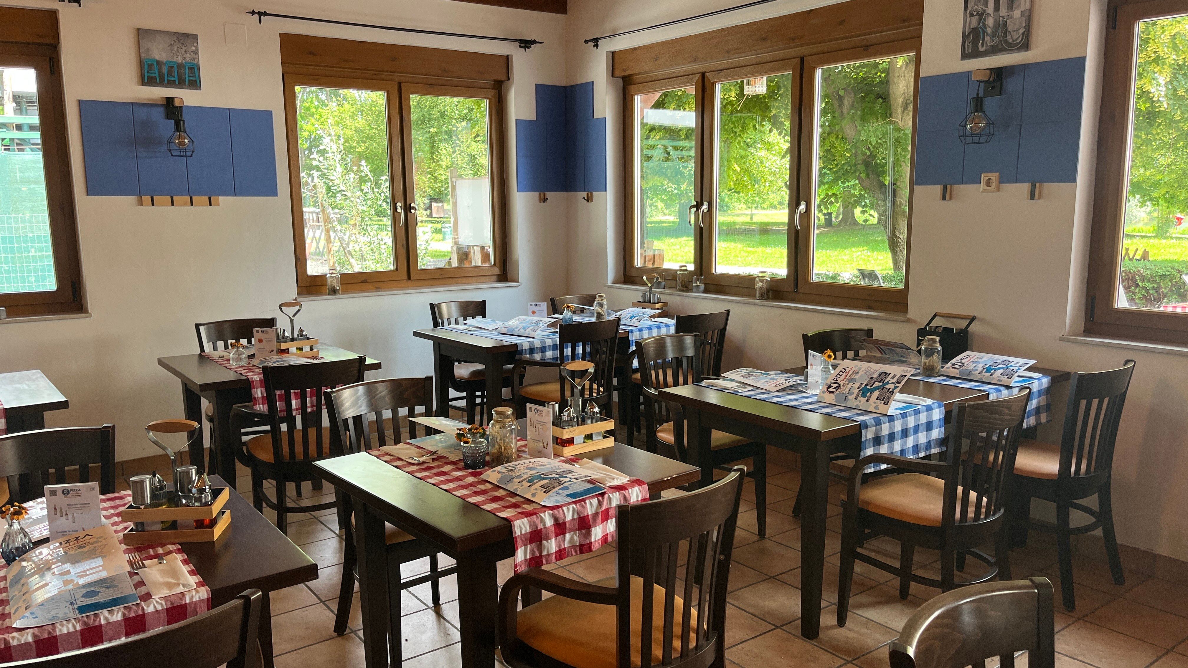 Interior view of the pizzeria. Black furniture with red tablecloth and cutlery. The Aubad can be seen through the windows in the background,
