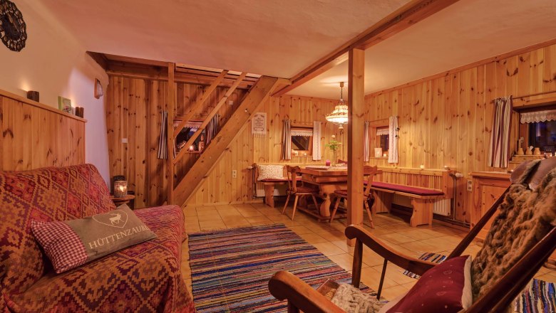 Cozy living room with wooden walls, sofa, rocking chair and dining table. Rustic ambience with carpet and warm light.