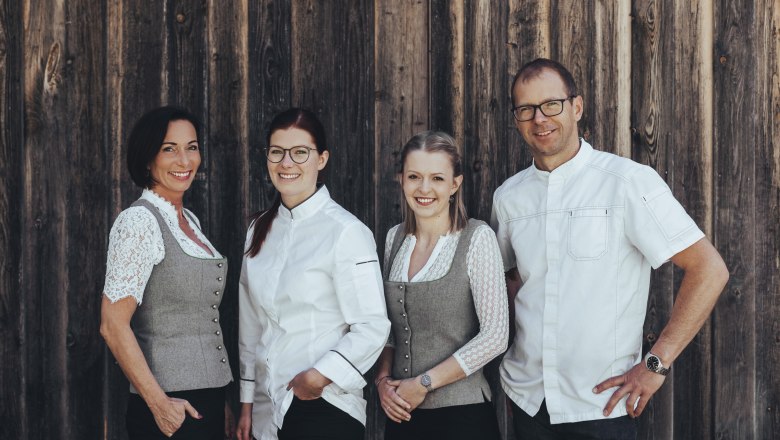 Four people are standing in front of a wooden wall, two women in traditional costume and two people in white clothing.