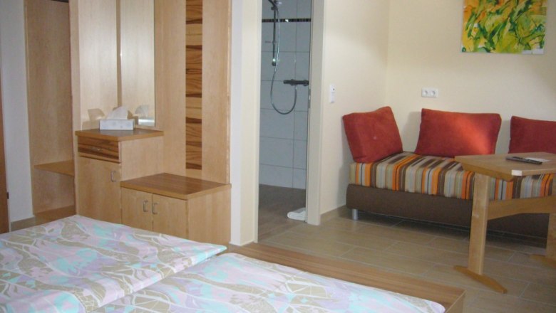 A cozy double room with two beds, a wardrobe, a small table and a couch. A bathroom can be seen in the background.