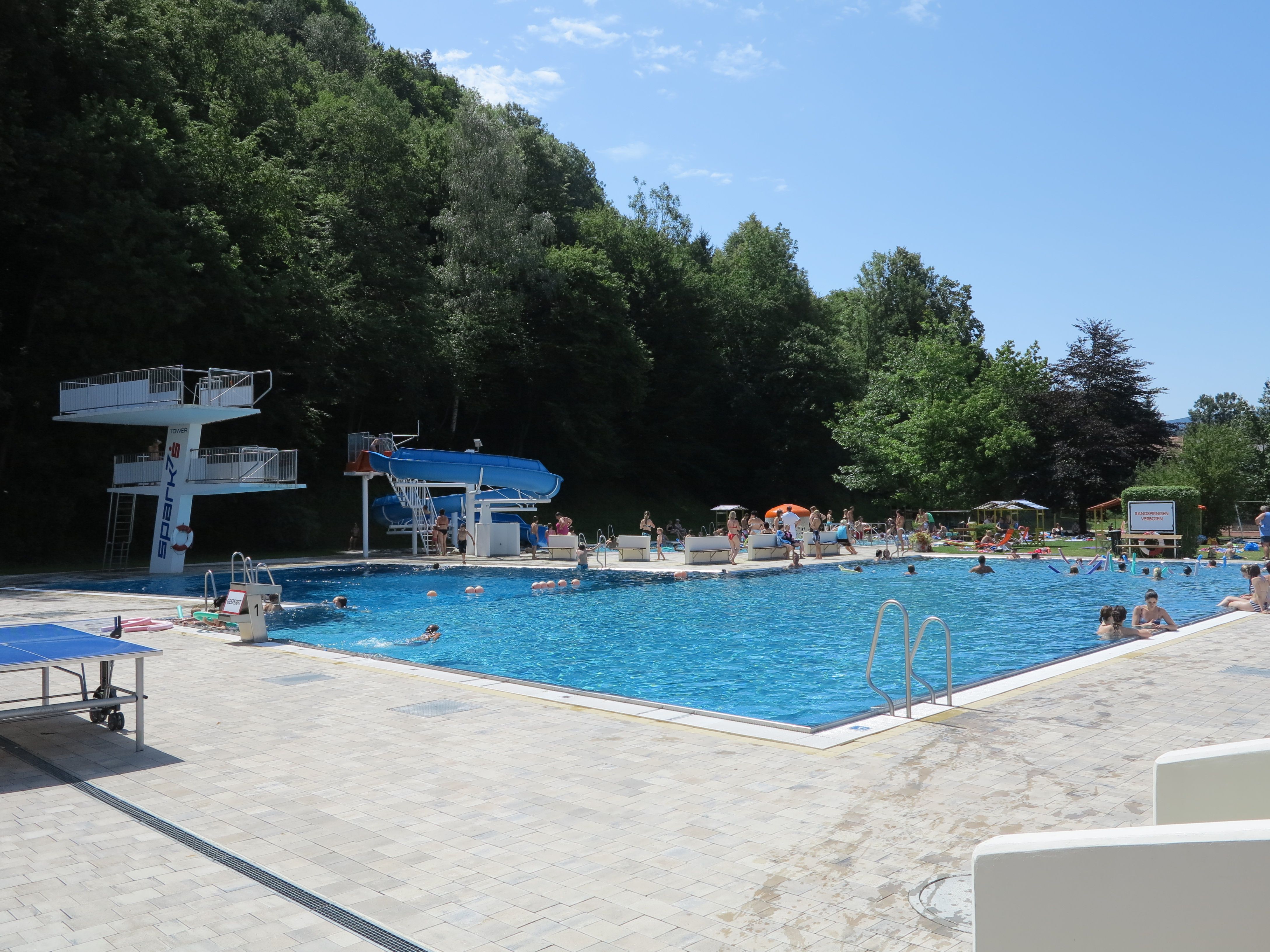 An outdoor pool with diving platform, slide and lots of bathers on a sunny day.