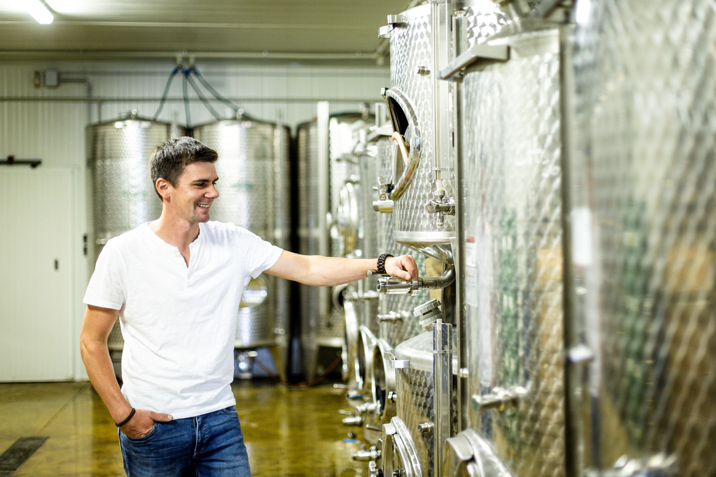 A man in a wine cellar stands smiling next to large stainless steel tanks.