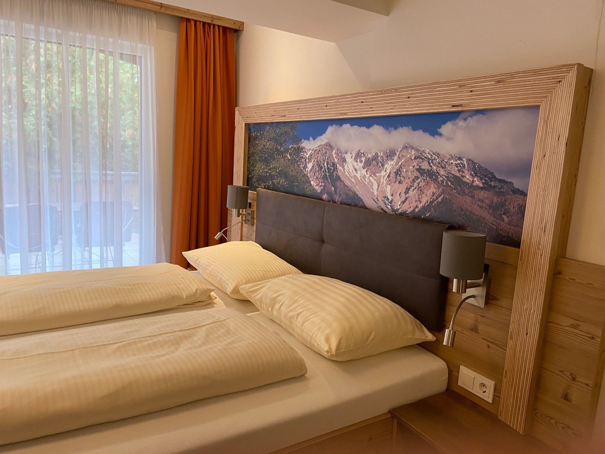 A cozy double room with a large bed, wooden furniture and a picture of mountains above the headboard. A window with curtains can be seen.