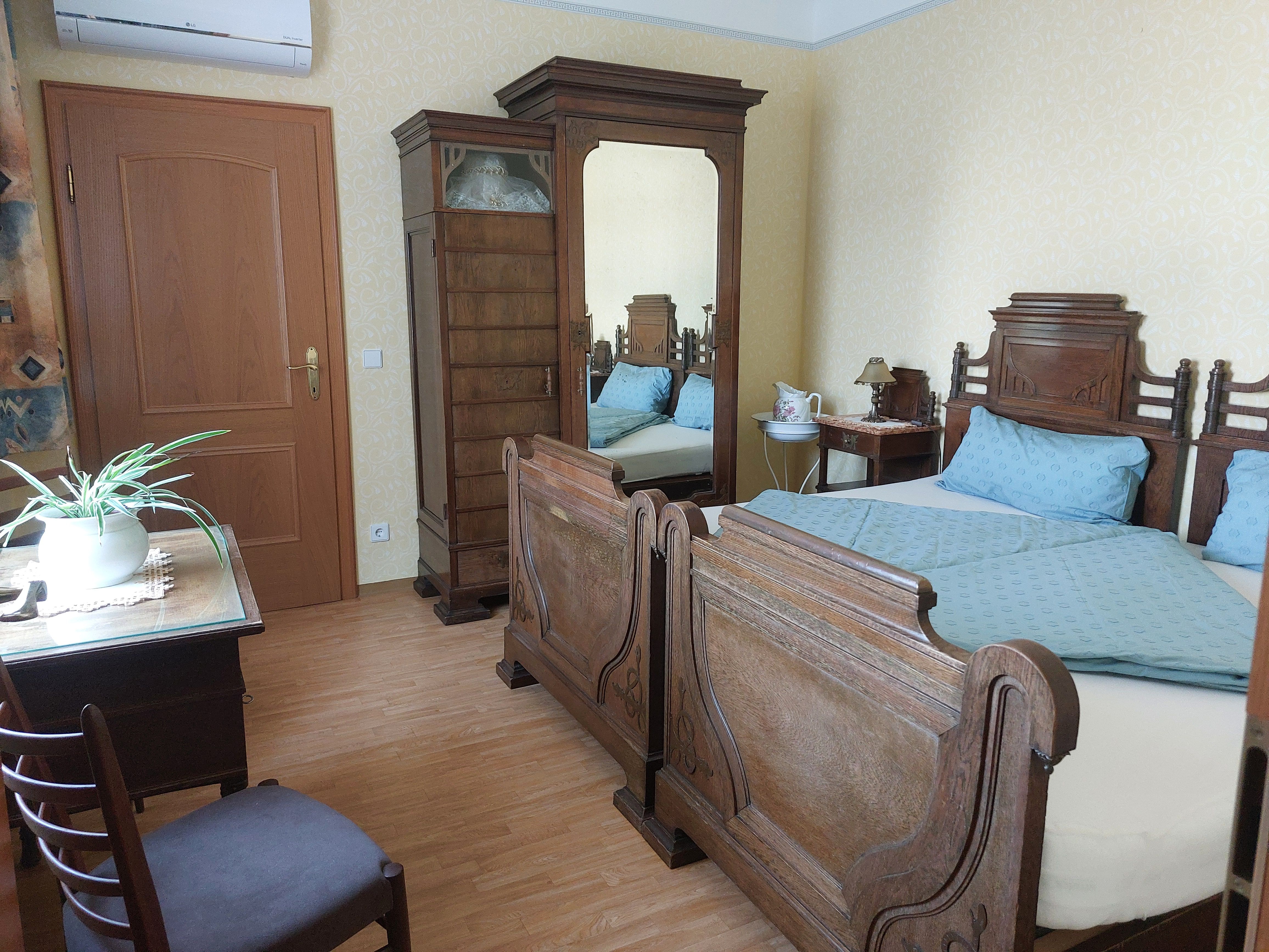 Bedroom with antique wooden furniture, double bed, wardrobe with mirror, table with plant and chair.