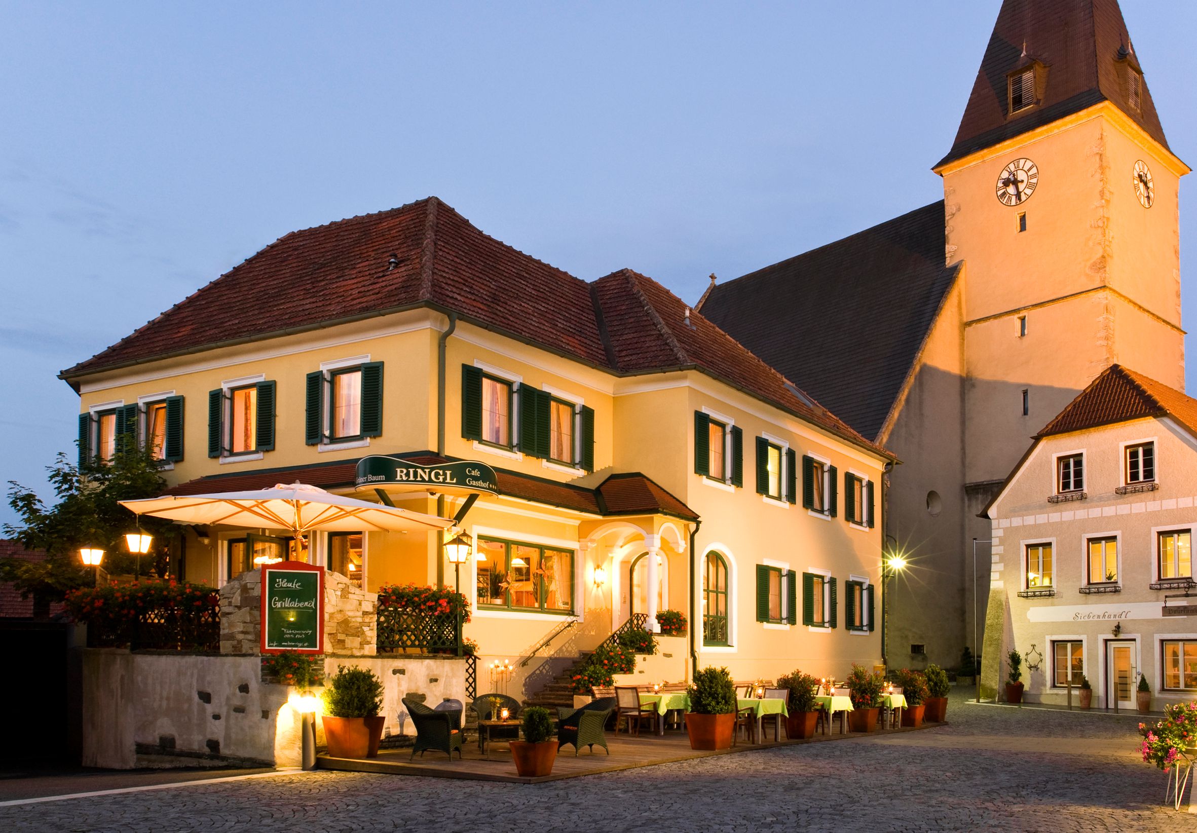 A traditional inn with a yellow façade and green shutters, illuminated at dusk, next to a church.