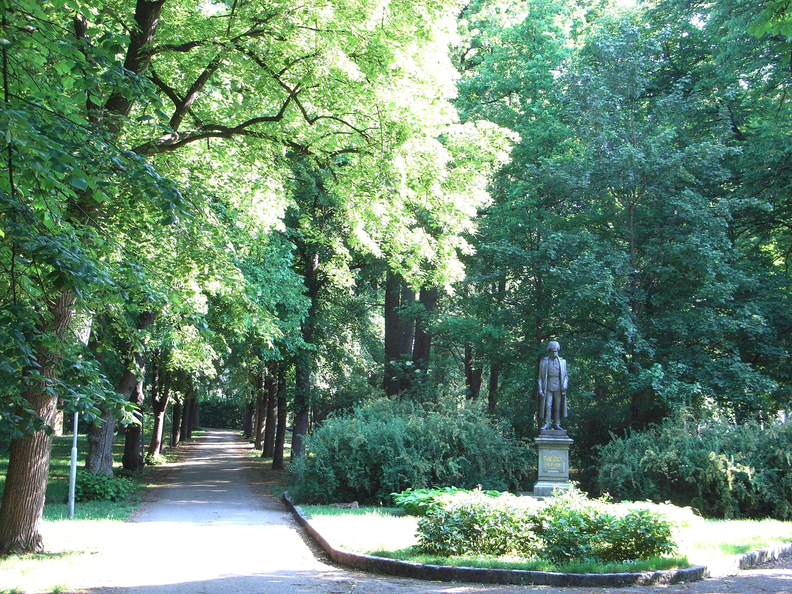 A sunny park path with a statue on the right, surrounded by lush greenery.
