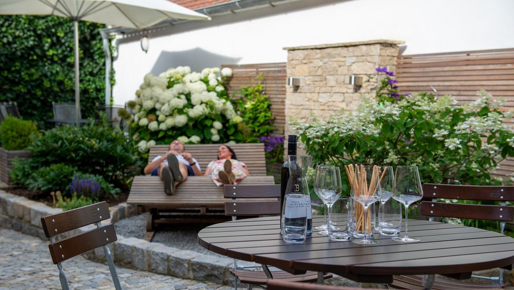 A table with wine glasses and a bottle, in the background two people on a lounger in the garden.