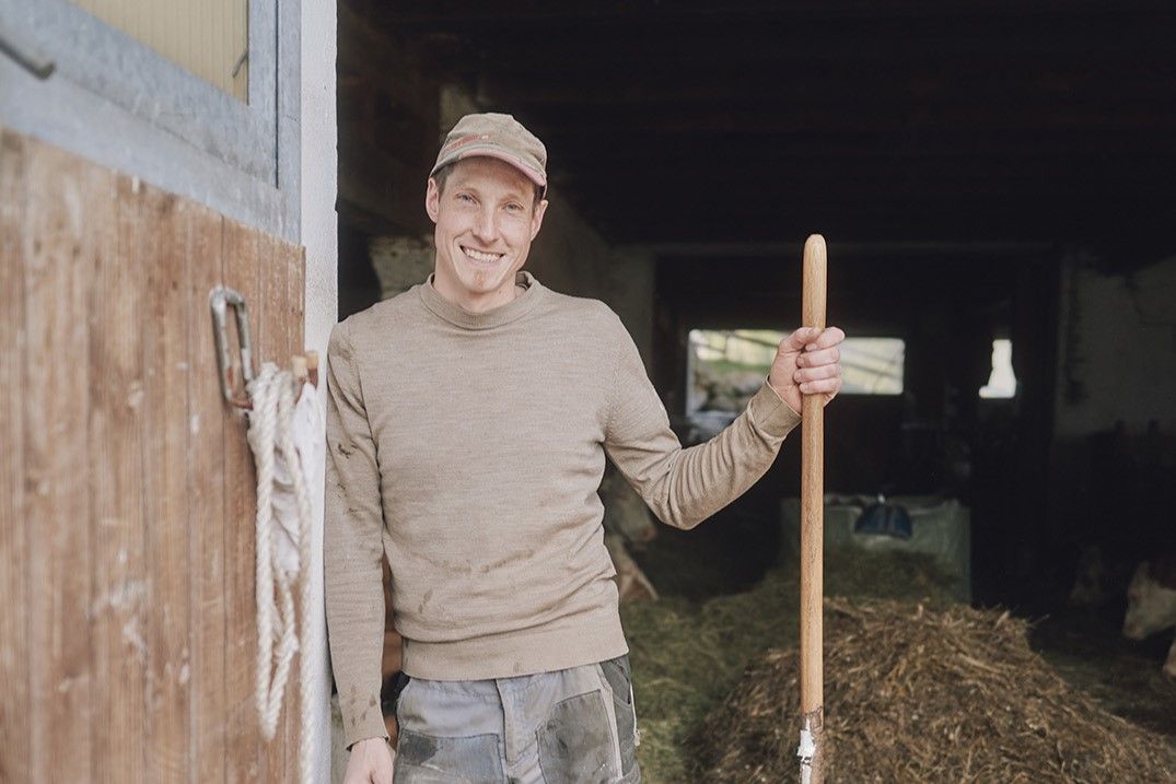 A smiling man in work clothes stands in a barn holding a pitchfork.