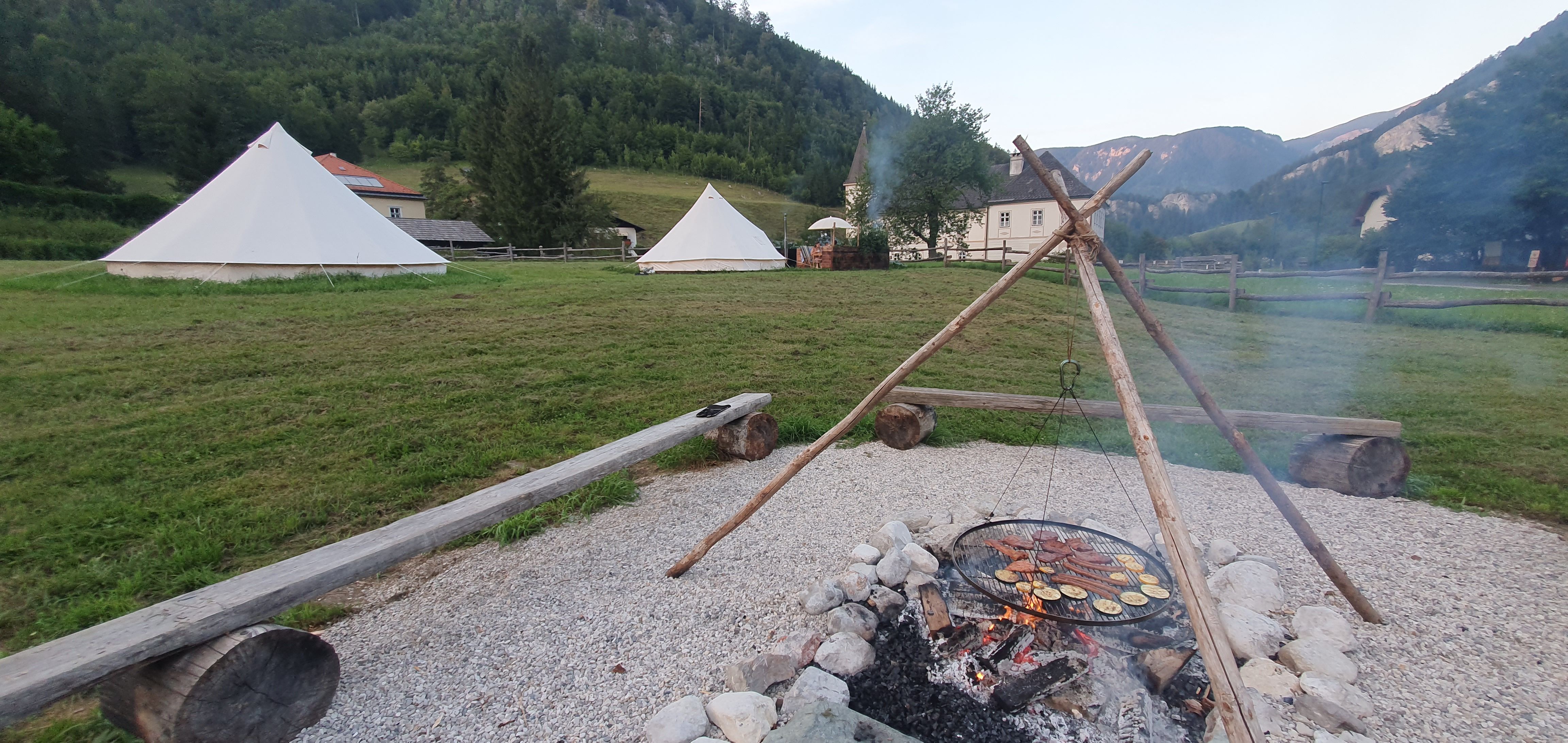 Campfire with barbecue over a fireplace, tents in the background, surrounded by mountains and meadows.