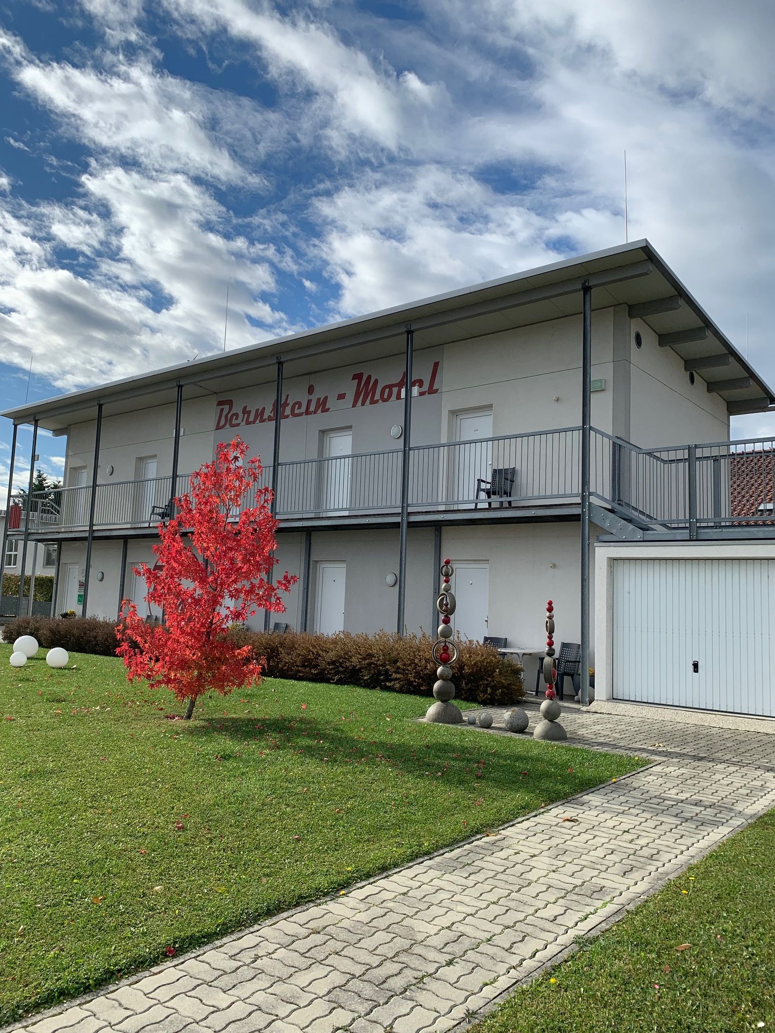 Two-story building with the inscription 'Bernstein Motel', red tree in the foreground, blue sky with clouds.
