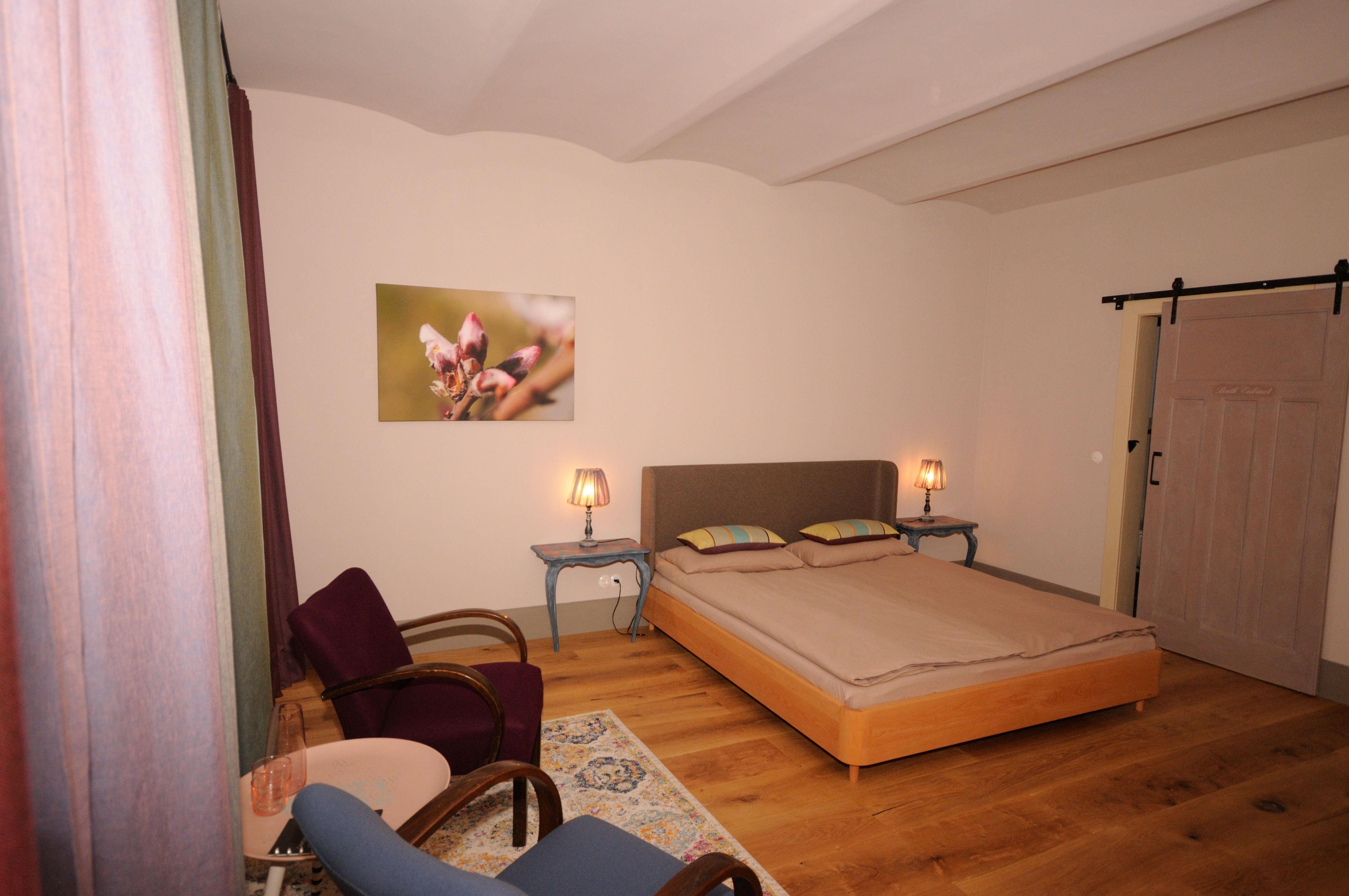 Cozy bedroom with double bed, two bedside tables with lamps, two armchairs and a picture on the wall.