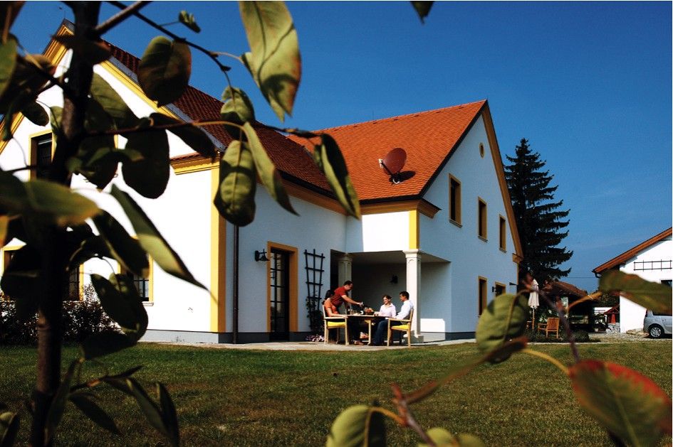 A white house with a red roof, surrounded by a green lawn and trees. People are sitting at a table outside.