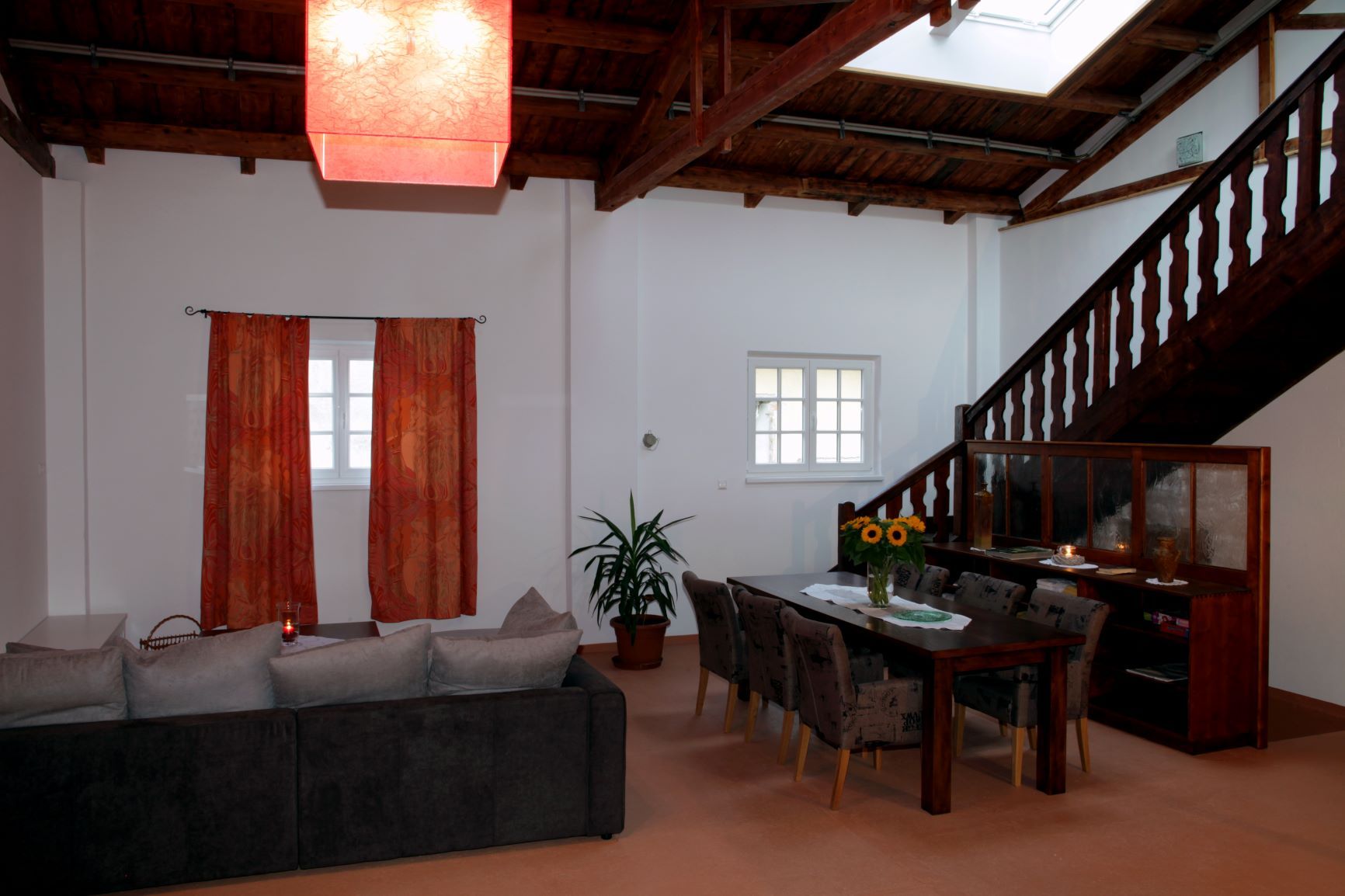 Living and dining area in a loft with wooden beamed ceiling, red lampshade, gray sofa, dining table with chairs and staircase.