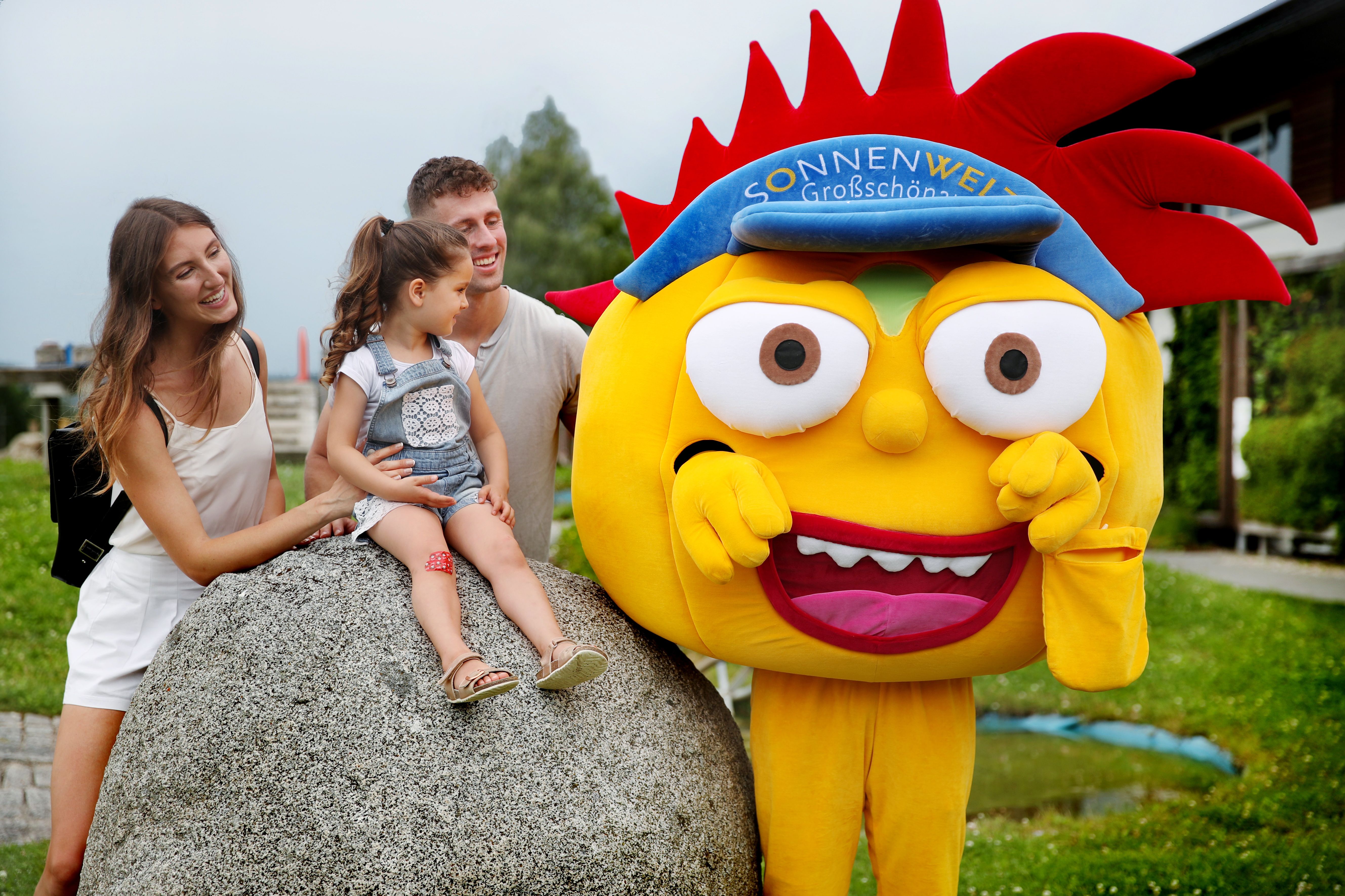 A family with a child sits on a large stone next to a yellow mascot with red hair and a blue cap outside.
