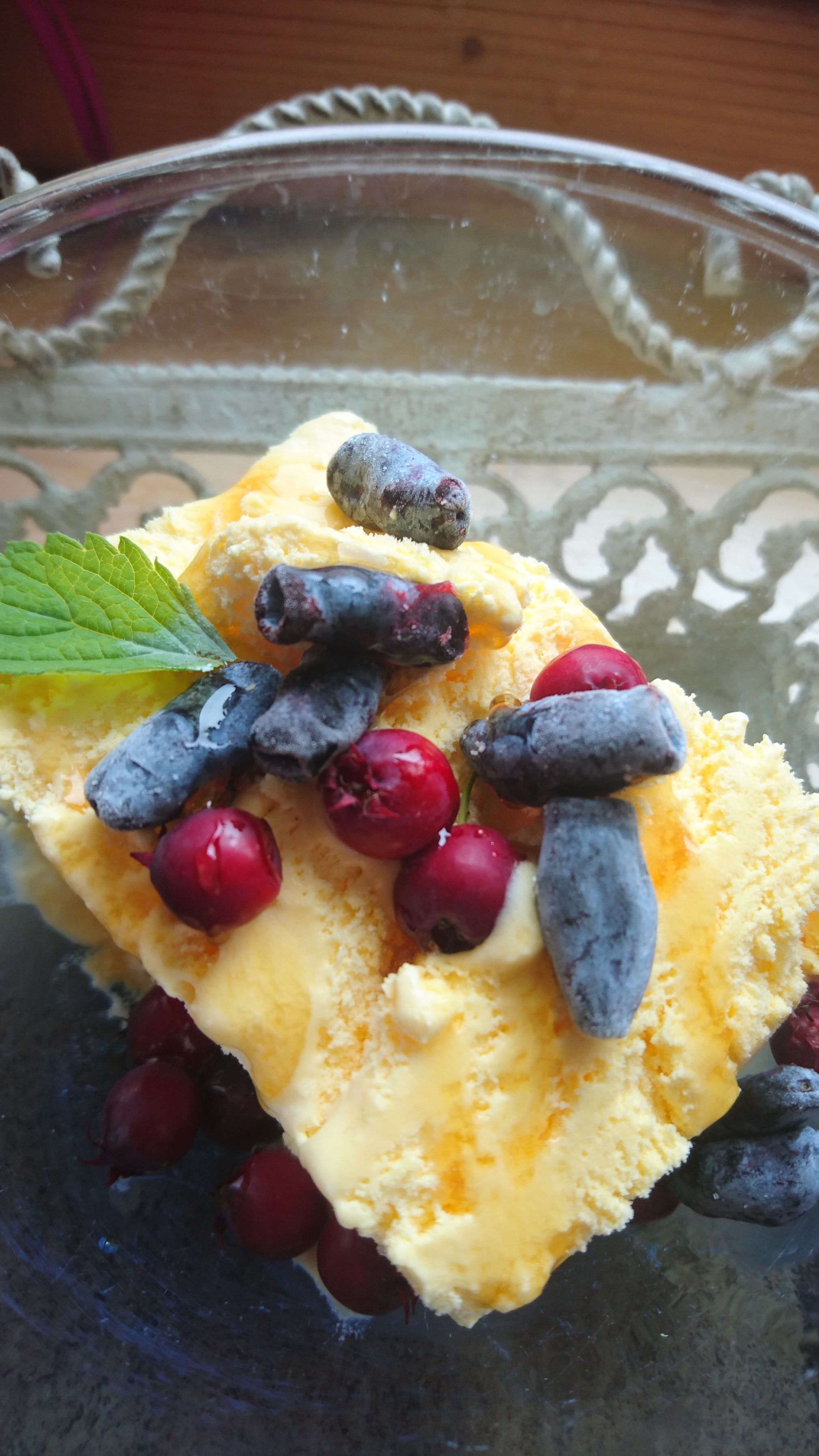 A dessert with yellow ice cream, red and blue berries, garnished with a green leaf, in a glass bowl.