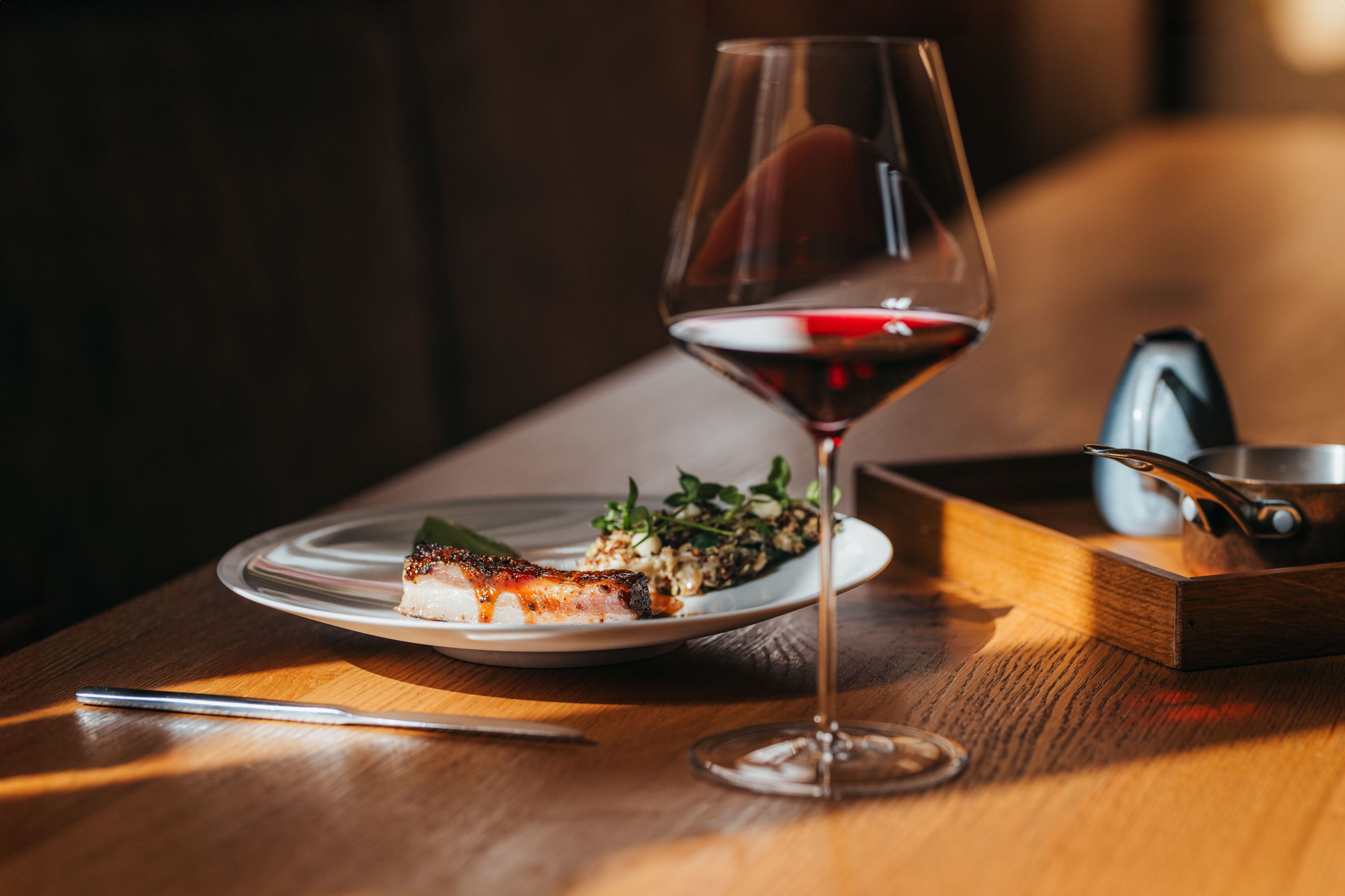 A plate of lamb belly, lettuce and wasabi cream next to a glass of red wine on a wooden table.