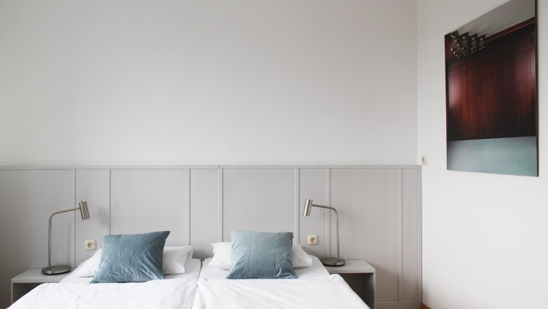 A simple guest room with a double bed, blue cushions, white walls and a picture on the wall.