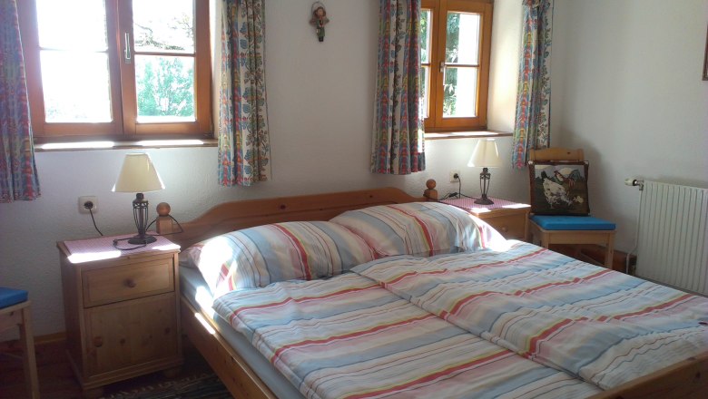 Cozy bedroom with double bed, striped bed linen, two bedside tables with lamps and colorful curtains on the windows.