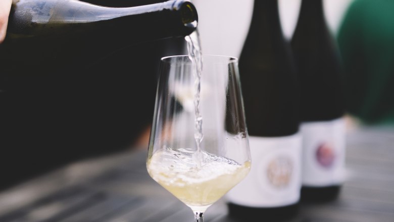 A glass of white wine is poured on a table, with two bottles of wine in the background.