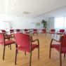 A bright seminar room with red chairs in a circle, a wooden floor and large windows.