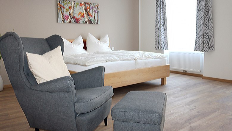 Gray, comfortable chair in the foreground. A double bed with white bed linen in the background.