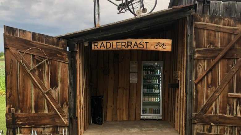 Wooden hut with 'Radlerrast' sign, bicycle on the roof, drinks inside.