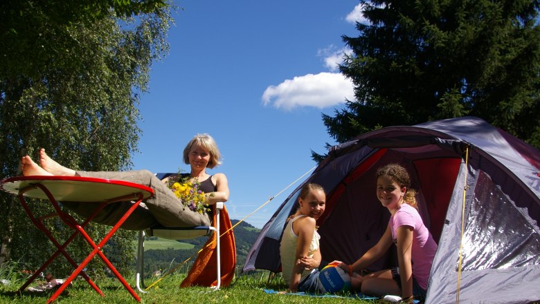 A woman relaxes on a camping chair next to a tent, while two children sit in the tent. Trees and blue sky can be seen in the background.