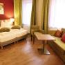 Cozy hotel room with double bed, green couch, table and chair. Warm colors and modern furnishings.