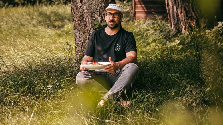 Man sitting in the grass under a tree holding a plate of food.
