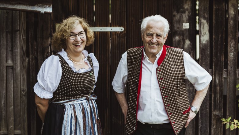 Two people in traditional costume in front of a wooden wall, smiling.