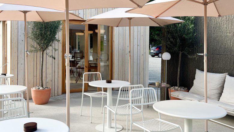 Modern terrace with white tables and chairs, parasols and wooden hut in the background.