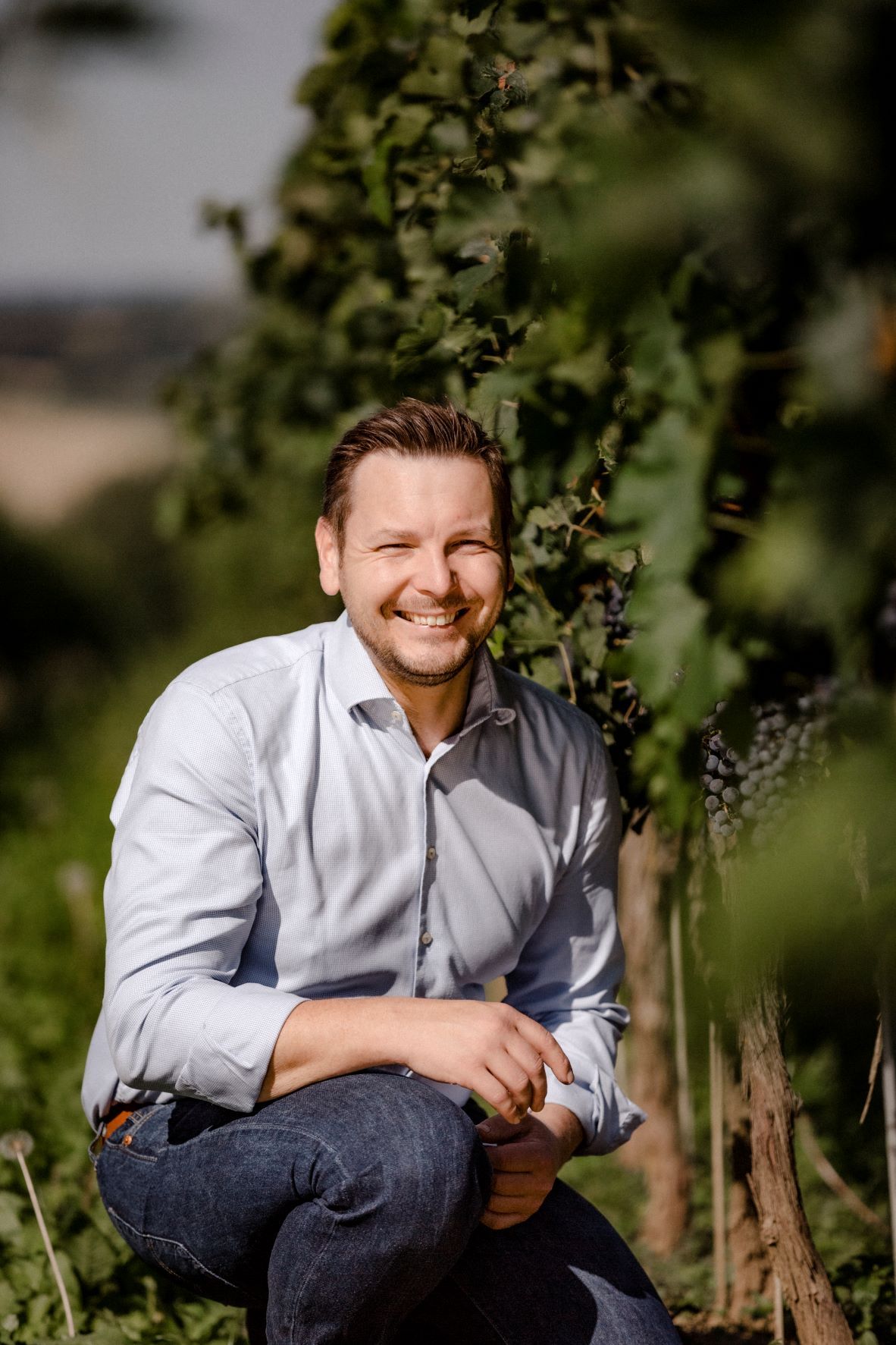 A man in a vineyard, smiling and squatting, wearing a light-colored shirt and jeans.