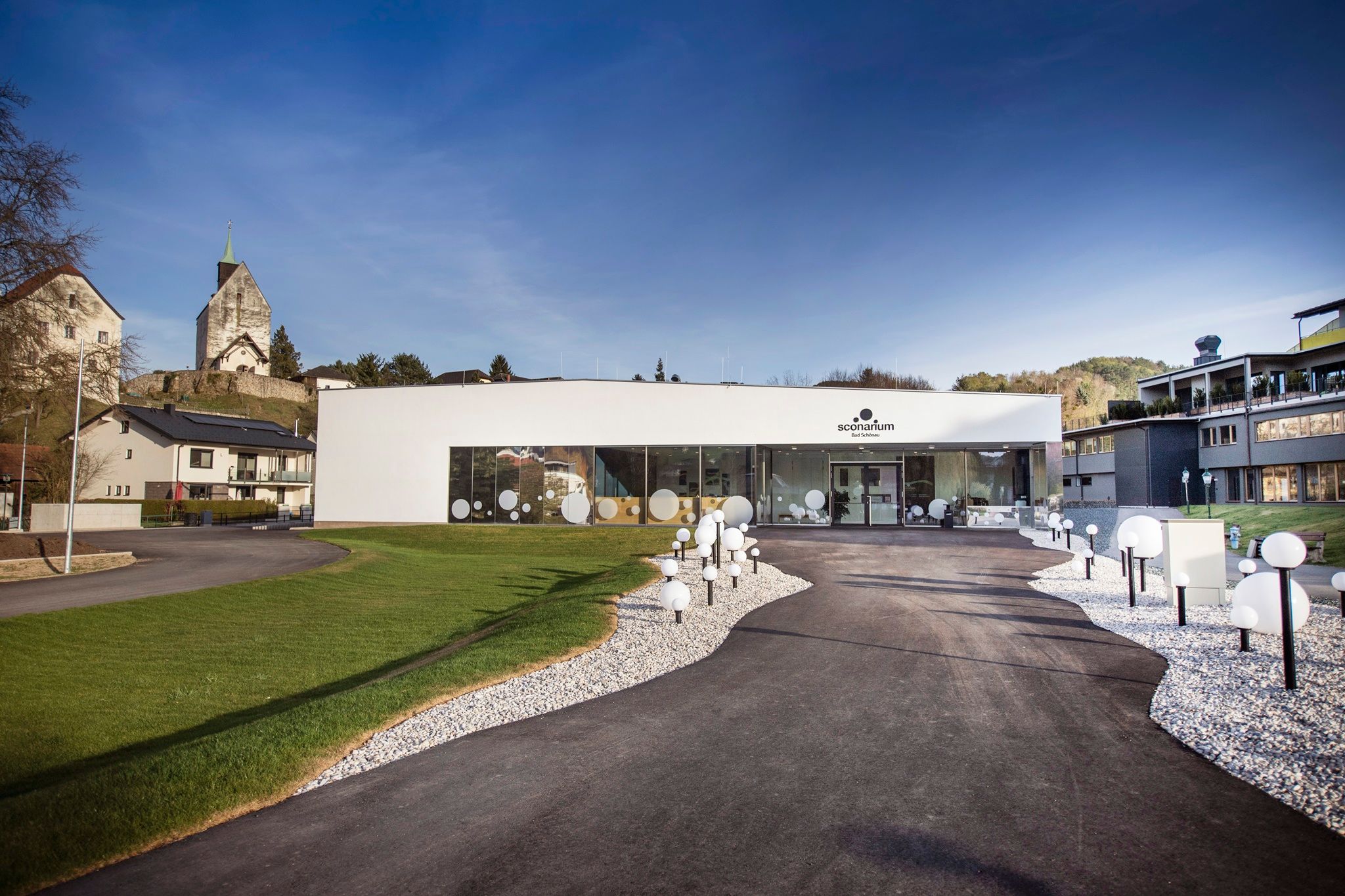 Modern Sconarium building with glass front and decorative globe lights, surrounded by green spaces and the fortified church in the background.