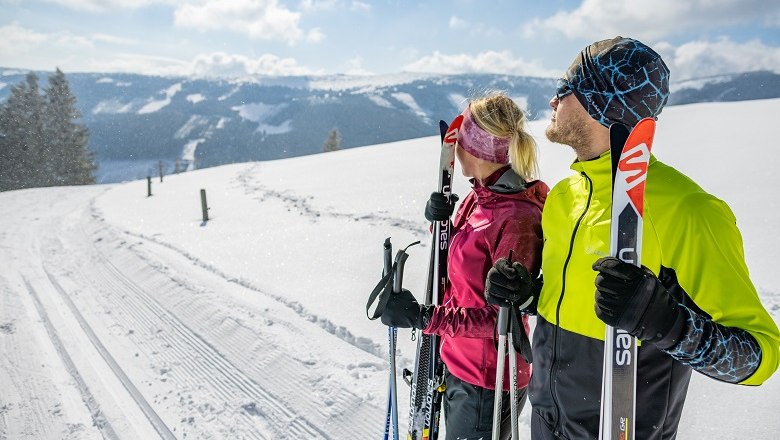 Two cross-country skiers on a snow-covered trail with a mountain panorama in the background.