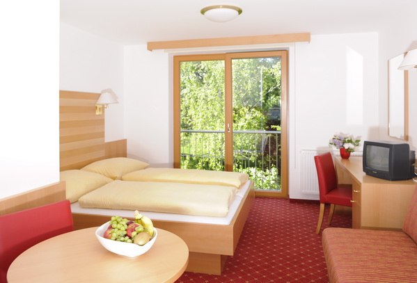 Bright hotel room with double bed, red carpet, table with fruit bowl and TV, view of green trees through a large window.